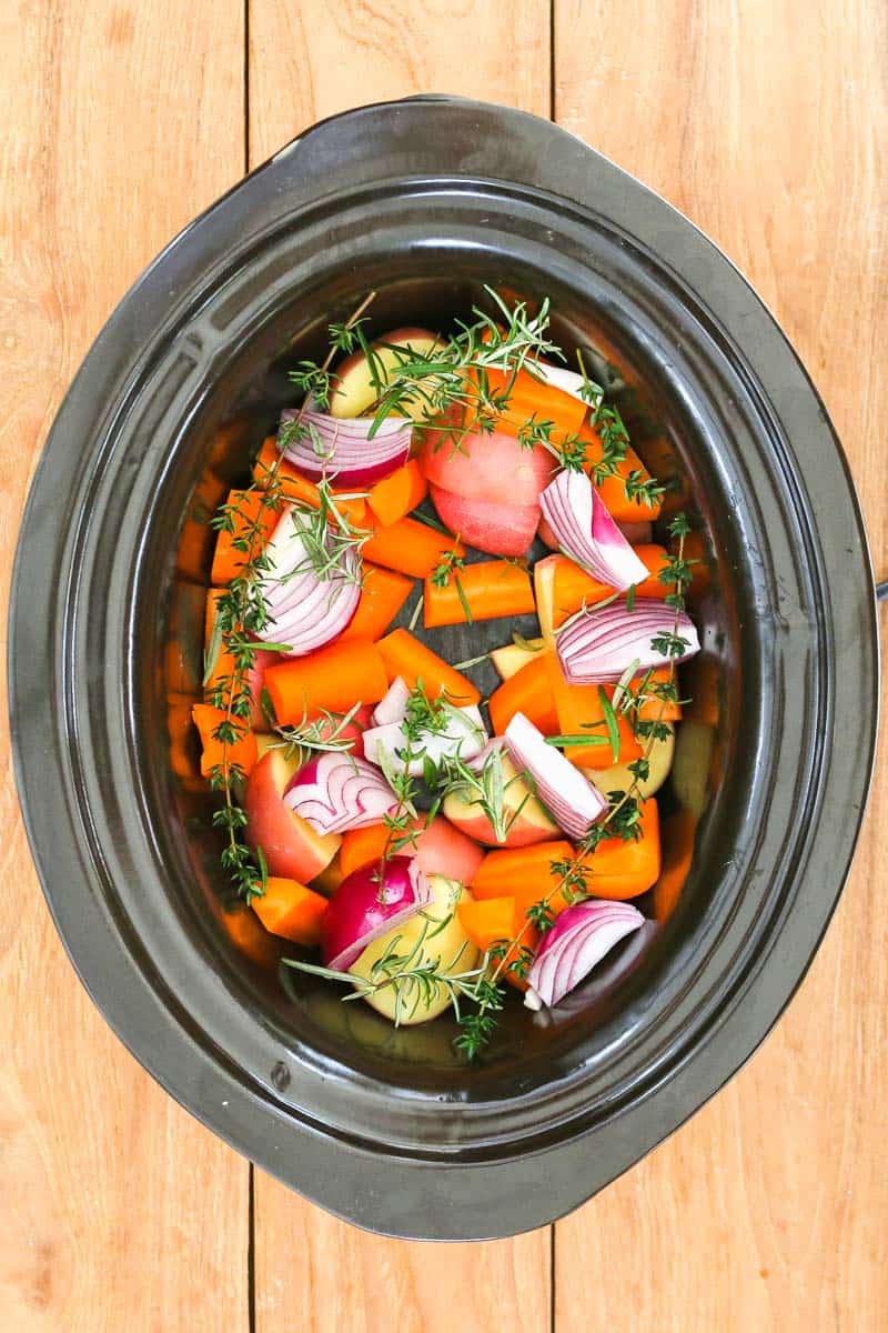 A crock pot filled with rosemary, thyme, potatoes, carrots and red onion. 