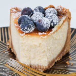 A slice of baked cheesecake decorated with blueberries and sprinkled with icing sugar on a plate.