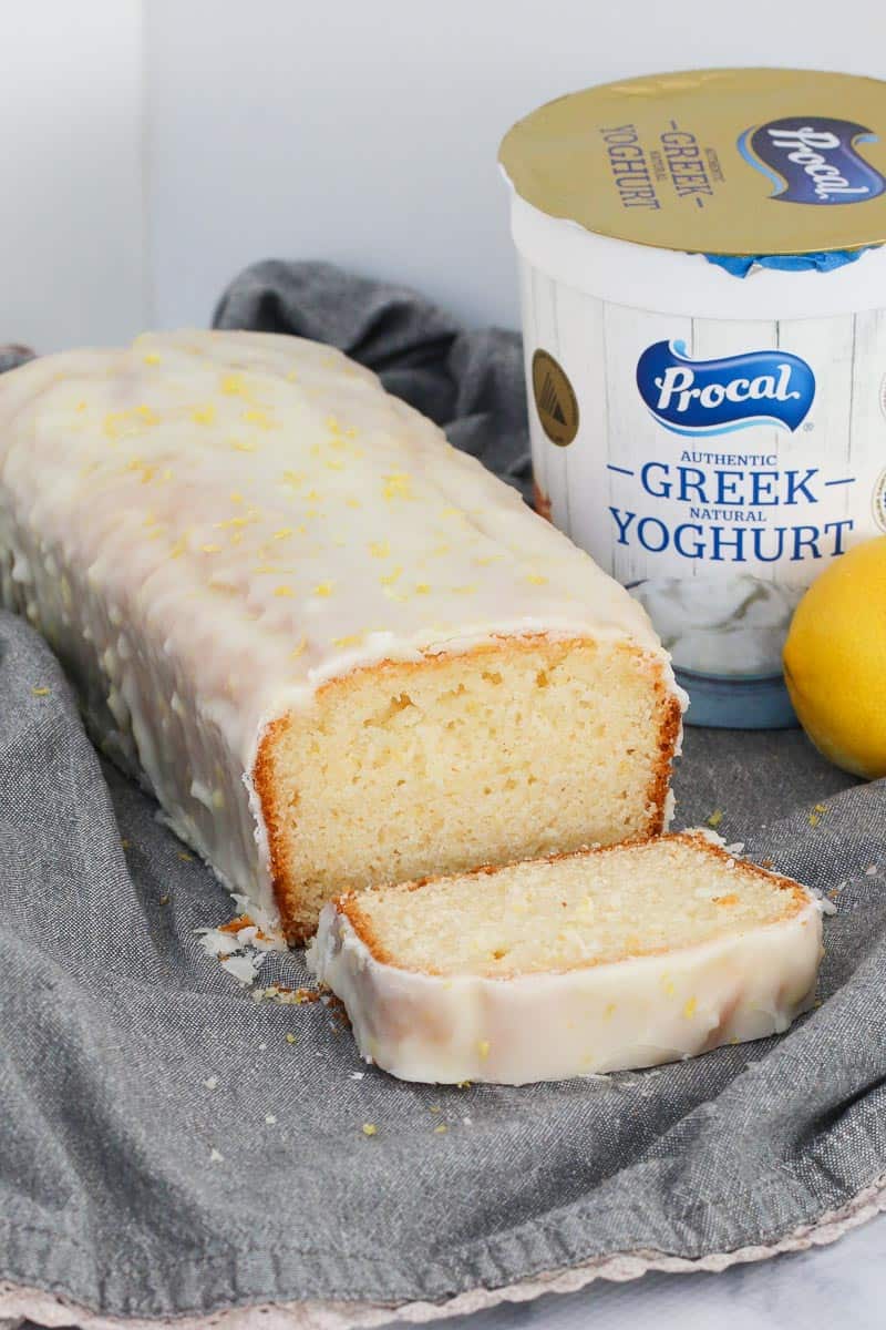 A tub of greek yoghurt behind a sweet cake with lemon icing. 