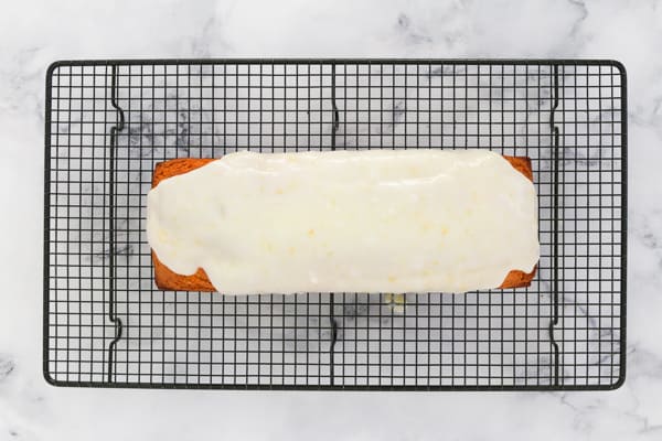 A long rectangular cake on a wire rack with a lemon glaze being poured over the top. 