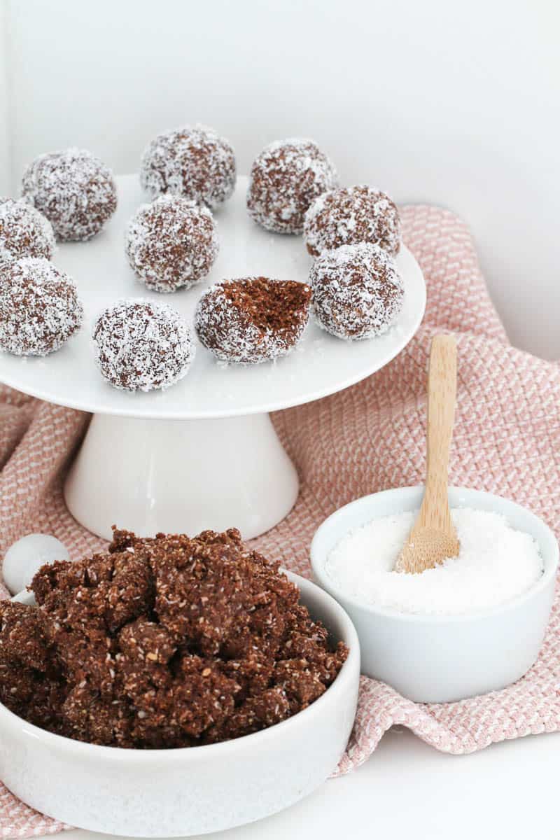 Rum balls with a half-eaten one on a white cake tray with a bowl of mixture and coconut in front.
