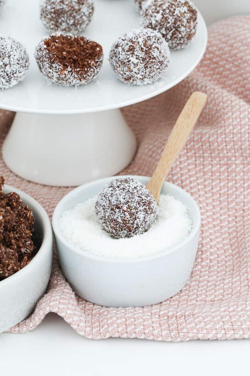 A white cake stand of rum balls on a pink tea towel with one ball being coated in coconut.