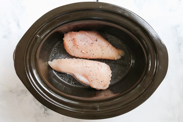 Two chicken breasts seasoned with salt and pepper in a slow cooker.