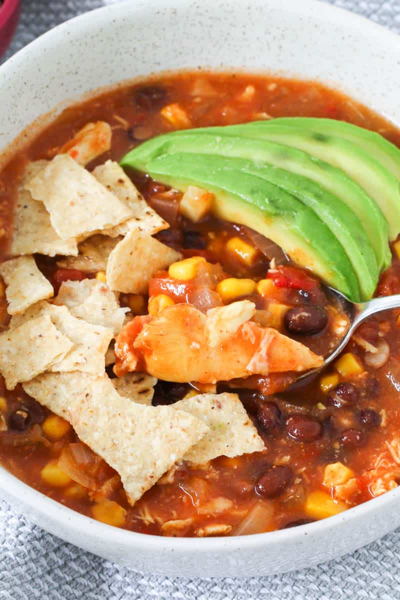 A spoonful of Mexican Chicken Soup made with beans and corn, and sliced avocado and tortilla chips on top