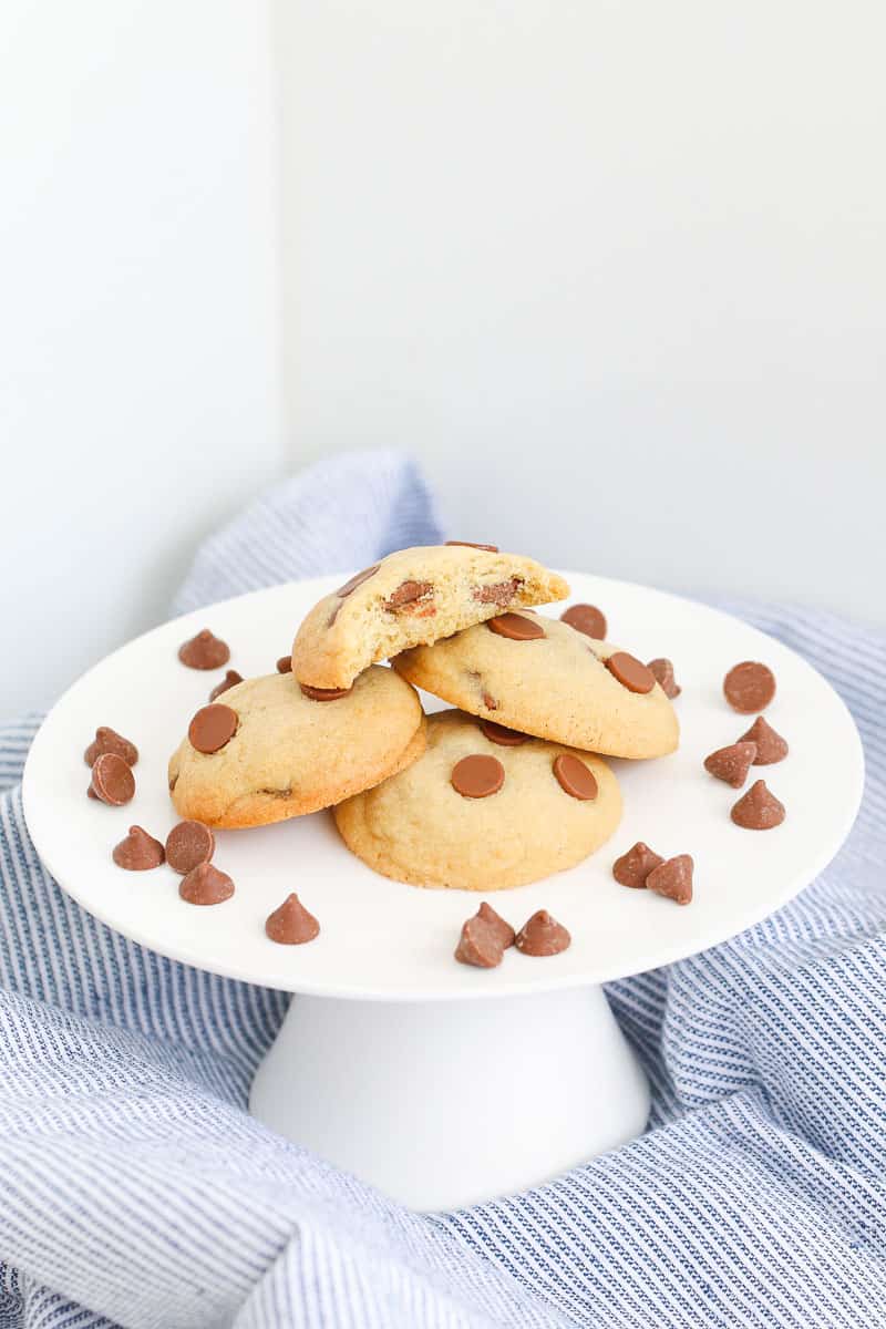 Chocolate chips around a pile of choc chip cookies on a white cake stand.