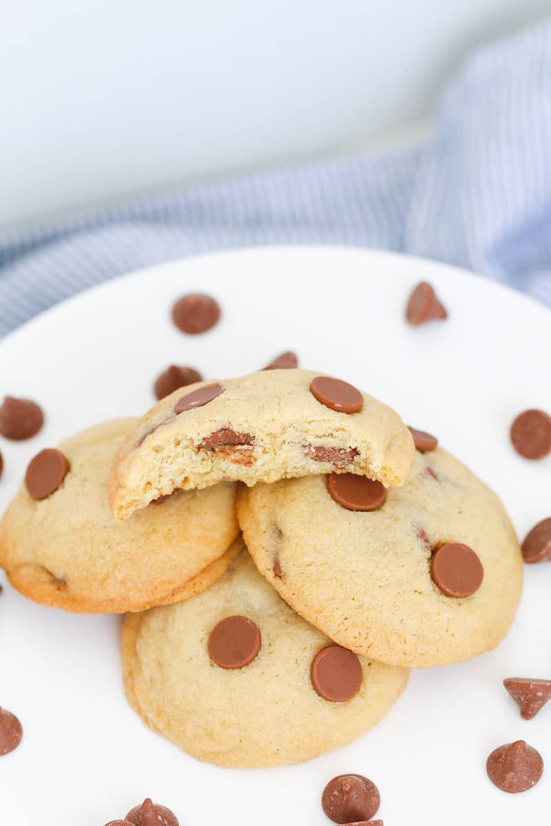 A half-eaten chocolate chip cookie on a pile of cookies with chocolate chips sprinkled around.