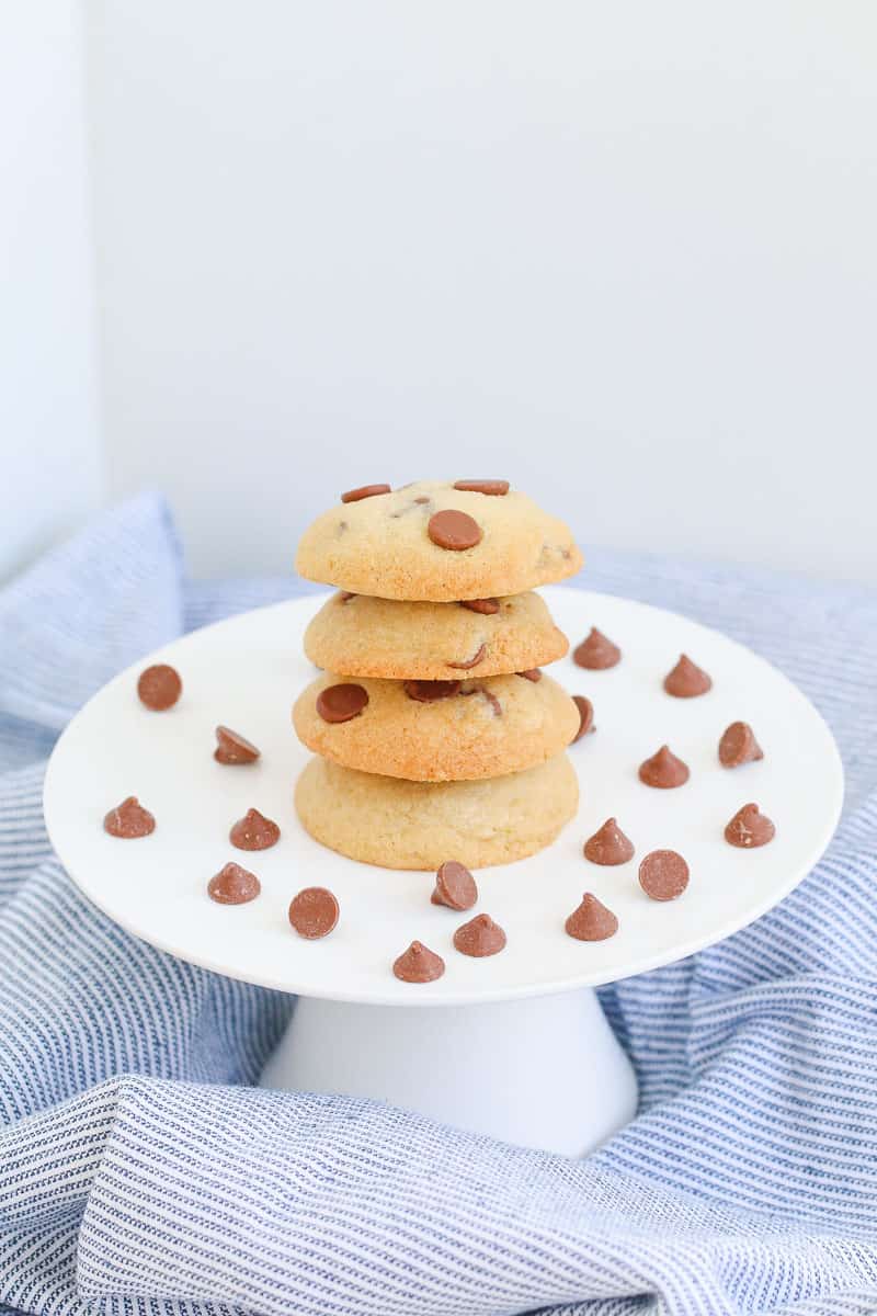 A stack of chocolate chip biscuits, with chocolate chips scattered around them on a white cake stand.