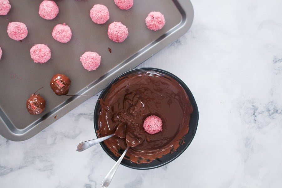 A pink cherry and coconut ball being dipped into a bowl of melted dark chocolate.