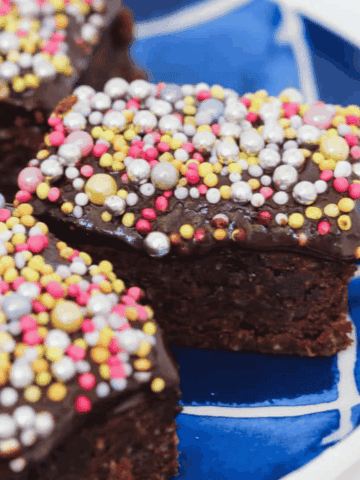 Chocolate slice topped with colorful sprinkles on a blue plate.ing a piece of chocolate slice with sprinkles.