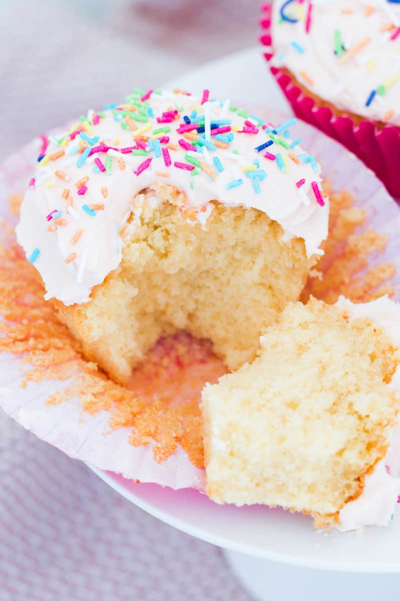 A fluffy vanilla cupcake with frosting and coloured 100s and 1000s broken in half.