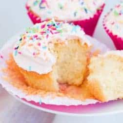 A cupcake with white icing and coloured sprinkles that has been cut and more cupcakes in the background.