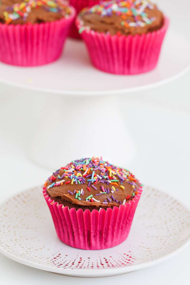 A pink cupcake case holding a chocolate cupcake with frosting and sprinkles.