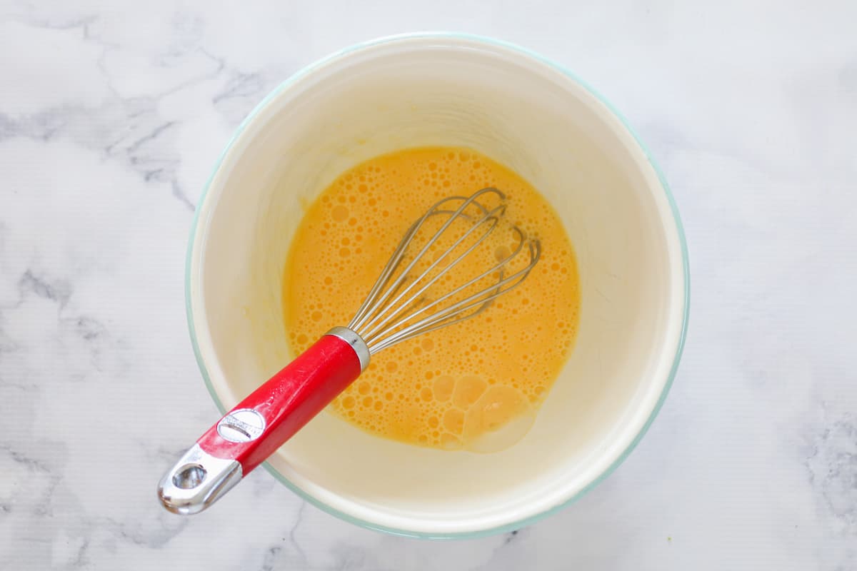 Beaten eggs in a mixing bowl with a red handled whisk.