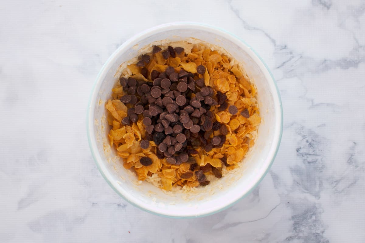 Chocolate chips, sultanas and cornflakes being added to a white mixing bowl.