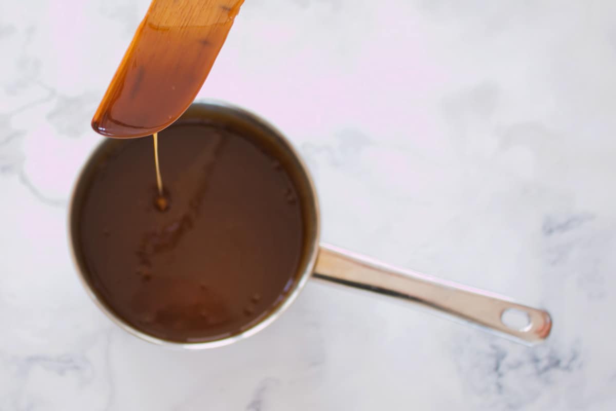 Caramel sauce dripping off a wooden spoon into a saucepan of caramel.