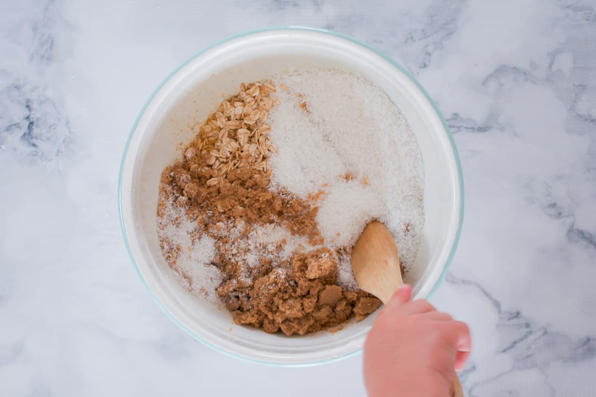 A hand mixing brown sugar, coconut and rolled oats in a bowl.