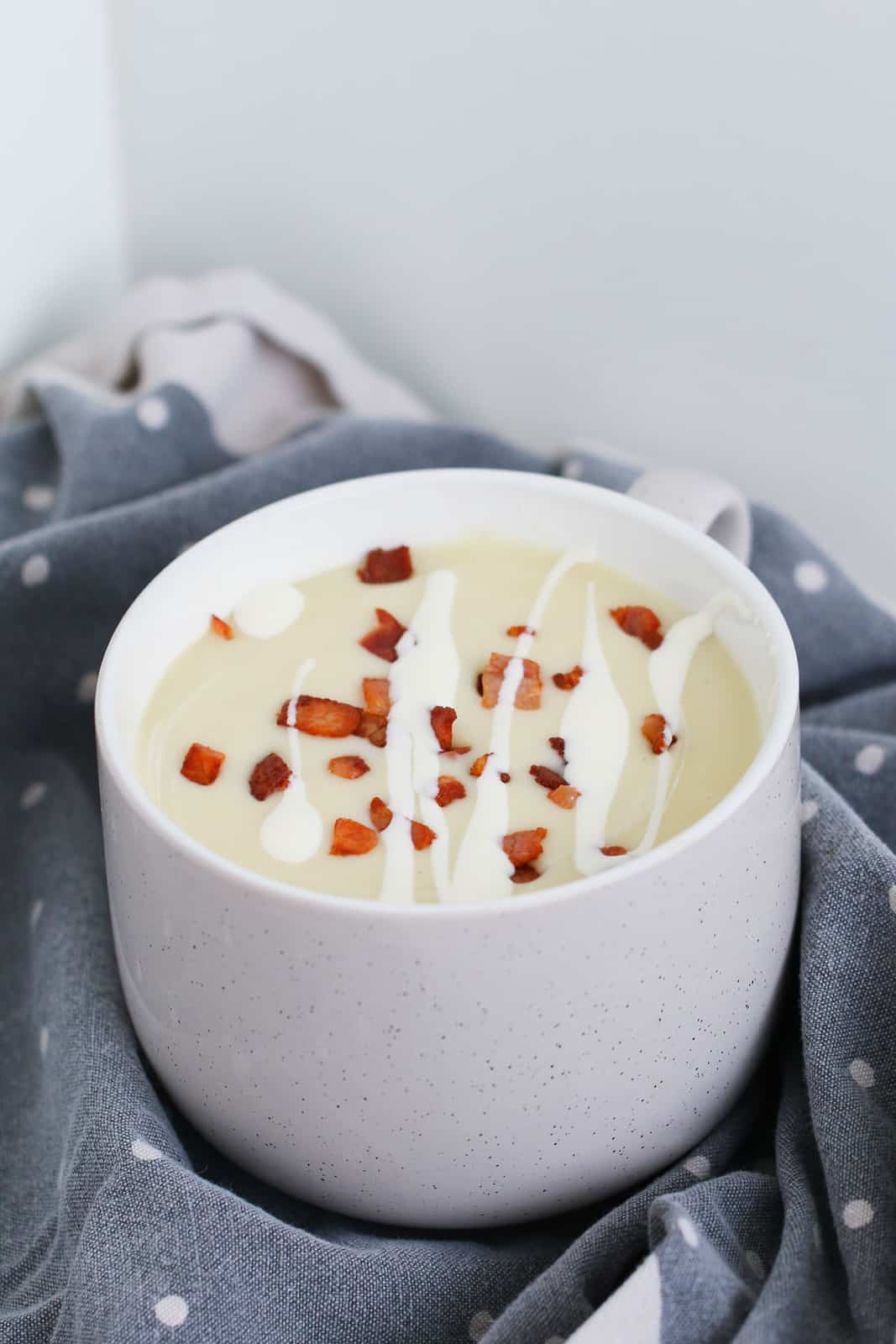 A mug filled with creamy vegetable soup and topped with bacon and cream.