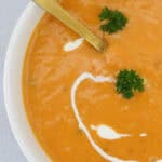A bowl of creamy roast pumpkin soup with a gold spoon and swirls of cream and fresh herbs.