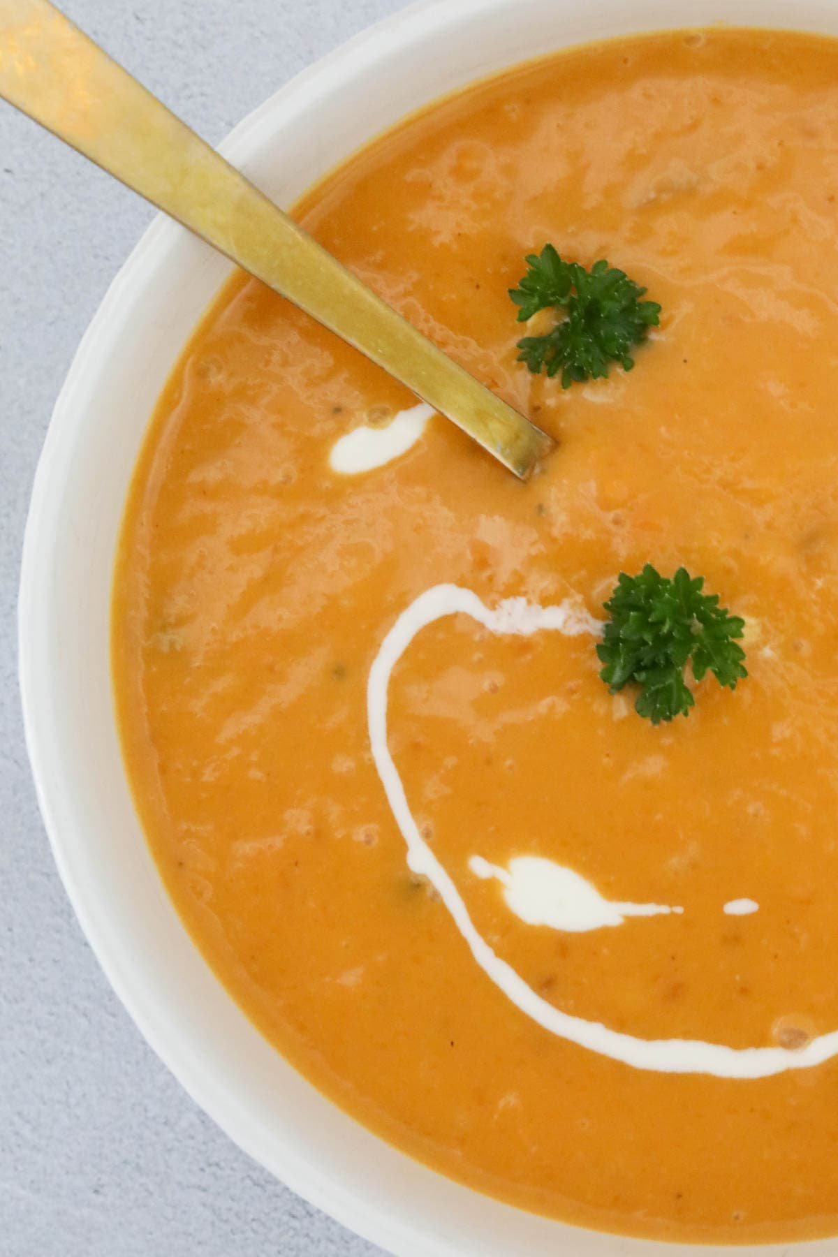 A bowl of creamy roast pumpkin soup with a gold spoon and swirls of cream and fresh herbs.