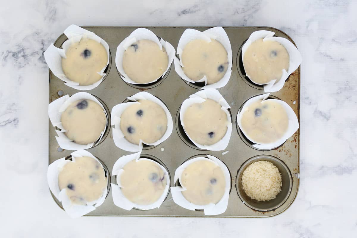 Muffin mixture with blueberries in white paper liners in a muffin tray.