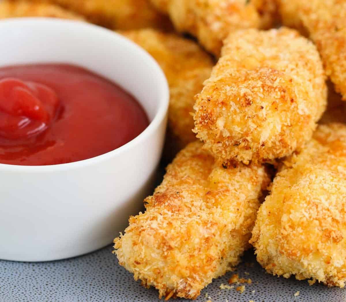 A stack of golden crunchy rectangular nuggets with red tomato sauce in a white bowl.