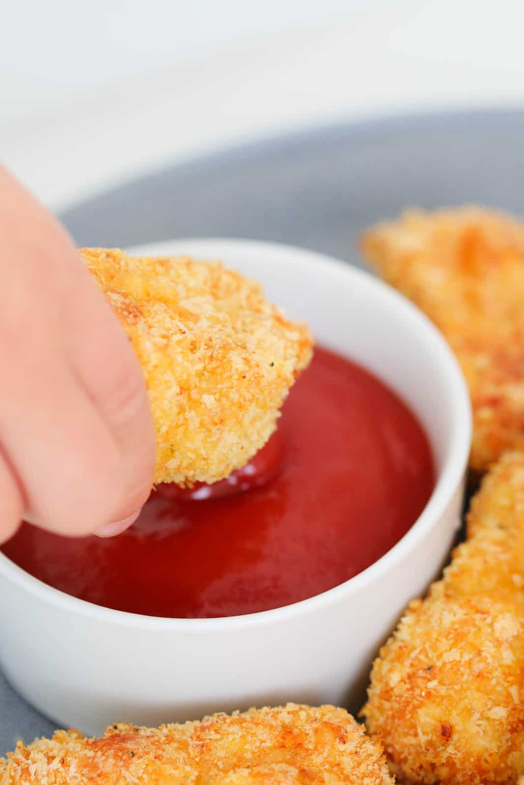 A hand dipping a crunchy crumbed homemade chicken nugget into a bowl of sauce.
