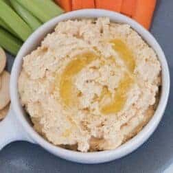 A bowl of hummus dip with olive oil drizzle and carrot and celery sticks in the background.