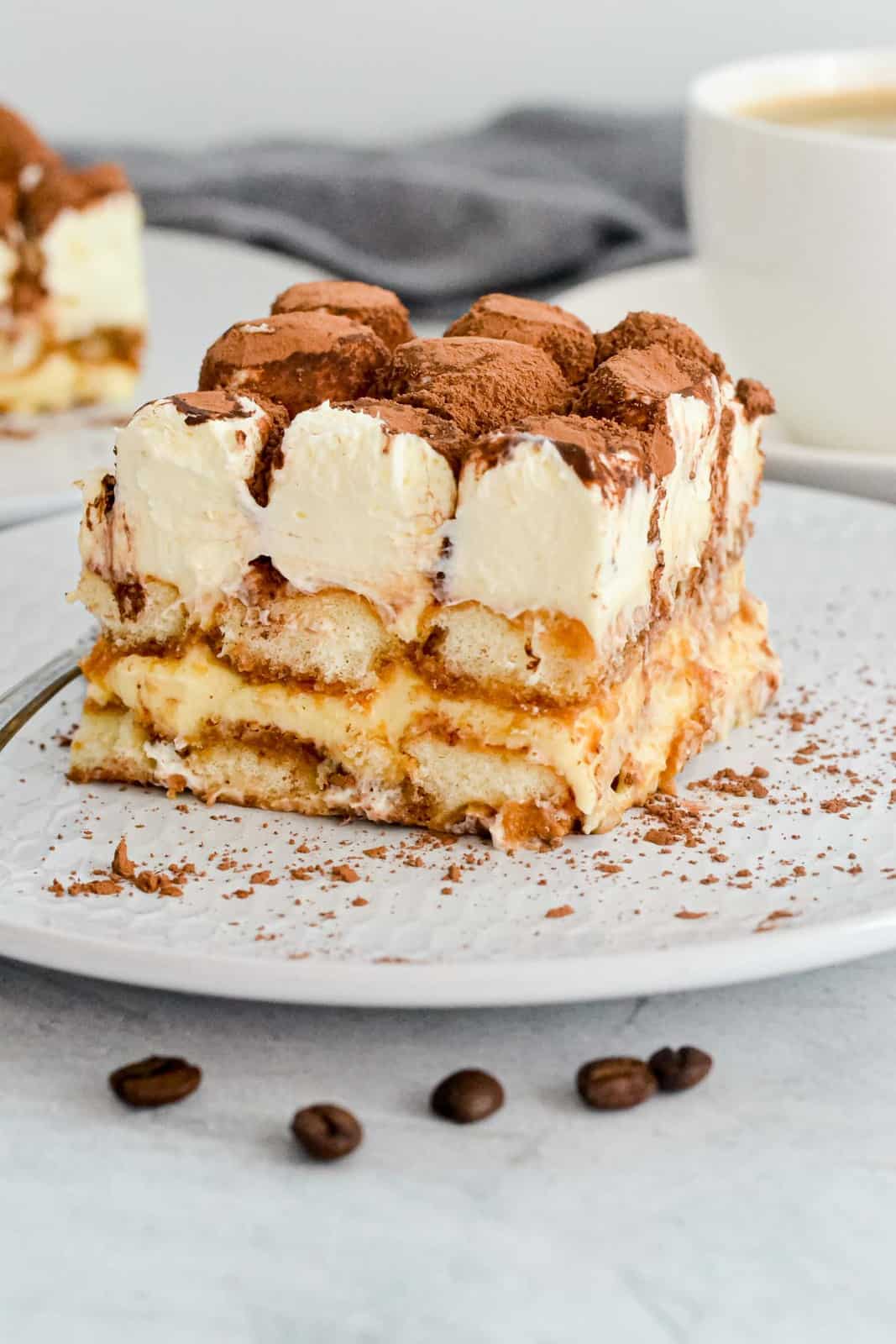 A piece of tiramisu on a plate with cocoa beans next to it.