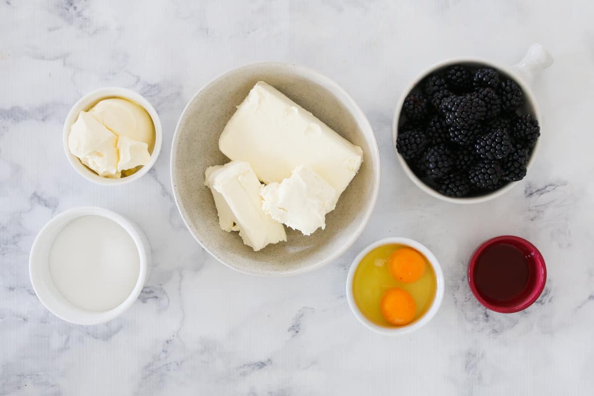 The ingredients for a blackberry cheesecake in bowls on a marble background.
