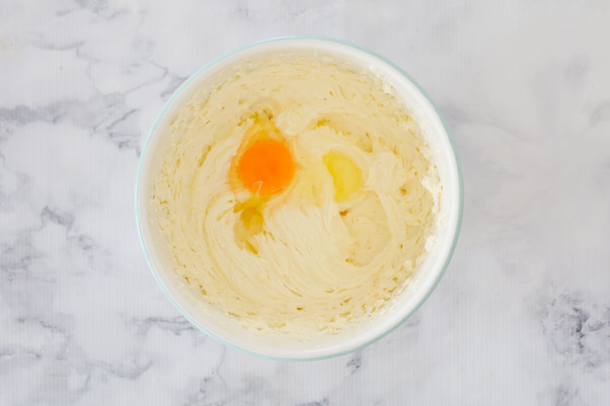 An egg in a white mixing bowl with beaten cream cheese and sour cream.