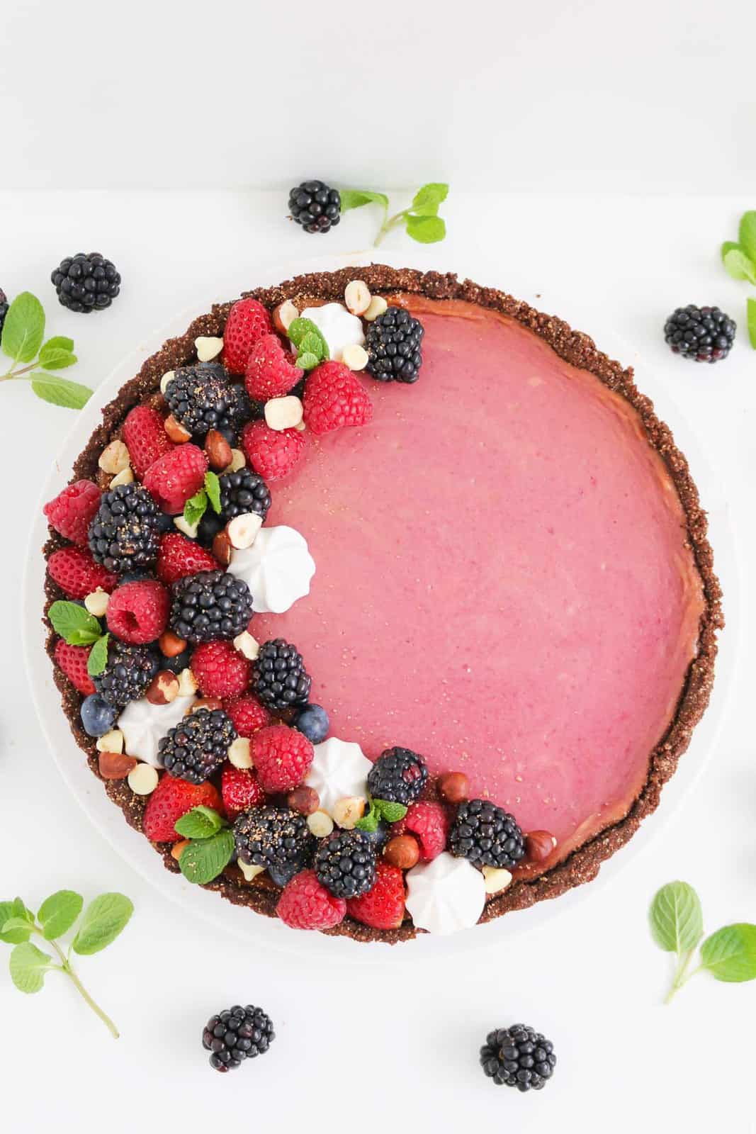 A pink cheesecake covered with berries, mint leaves and meringues with a chocolate crust.