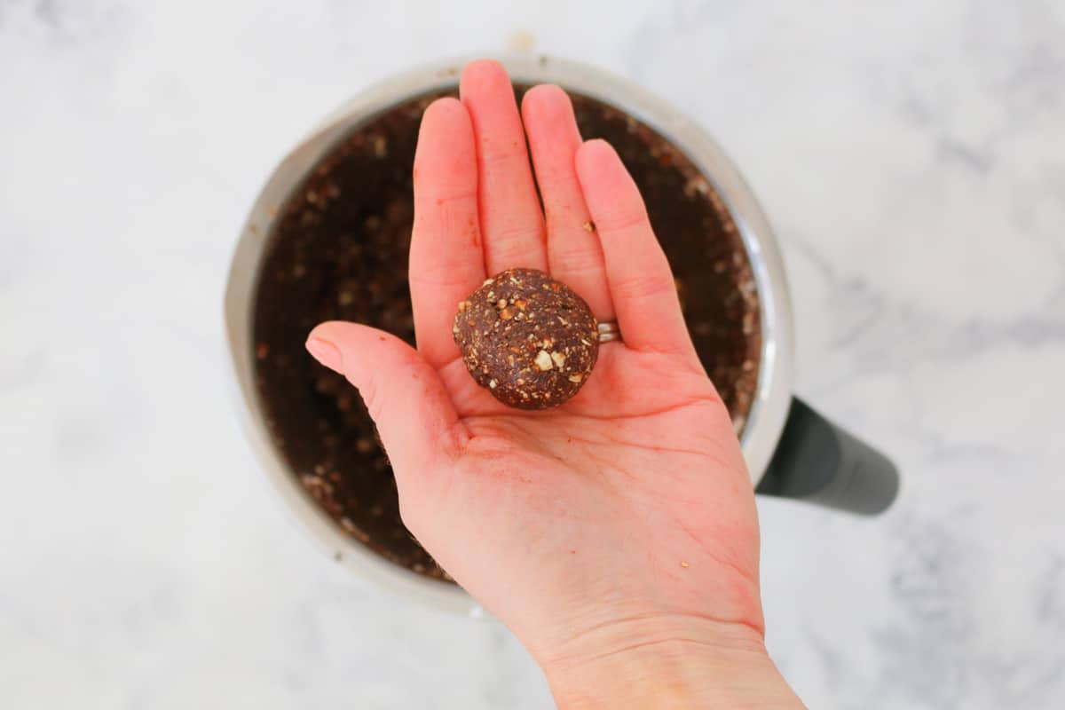 A hand holding a round date, almond and chocolate protein ball above a bowl of mixture.