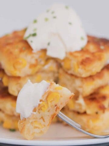 A fork with a piece of corn fritter with a stack of fritters in the background topped with sour cream.