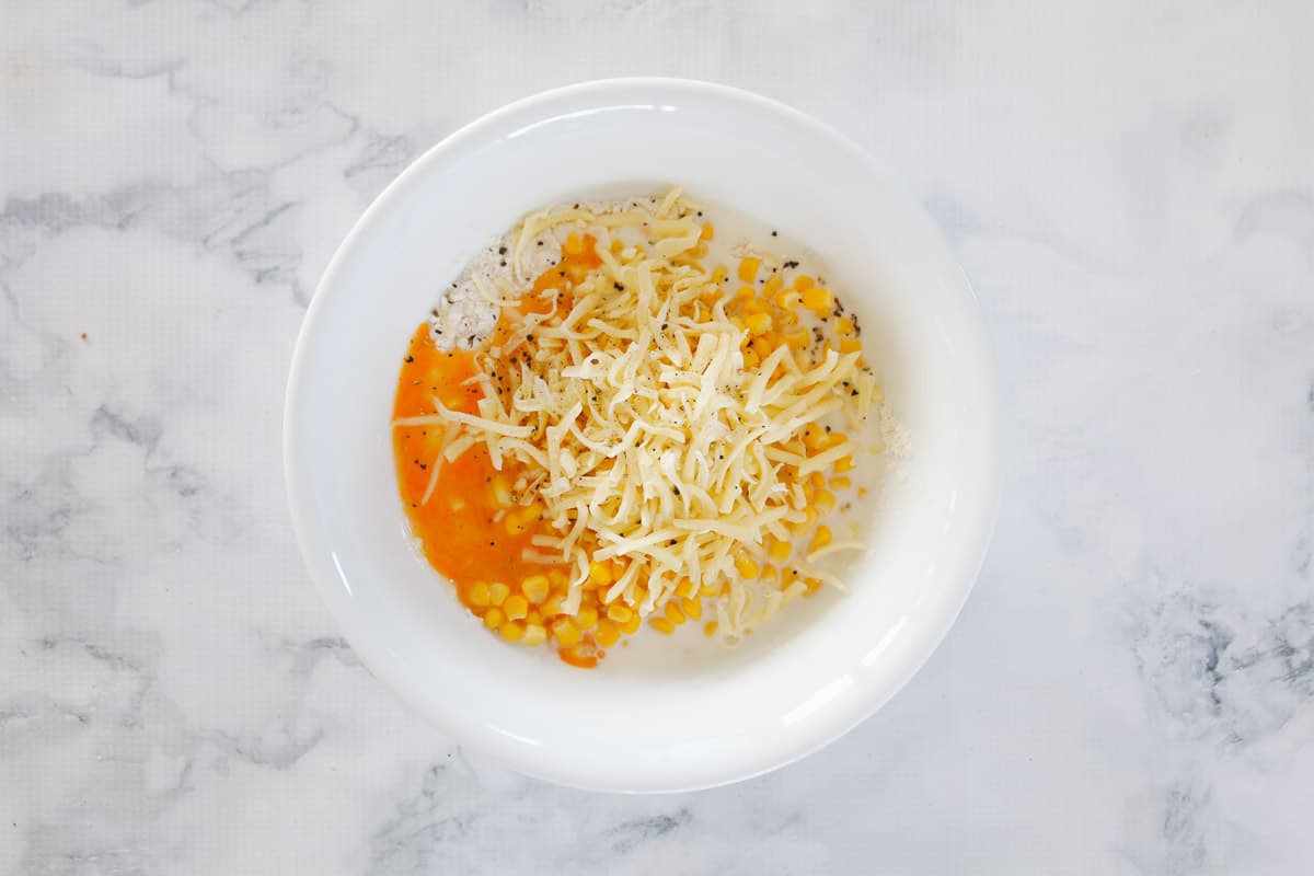 Grated cheese, eggs, milk, corn and flour in a white mixing bowl.