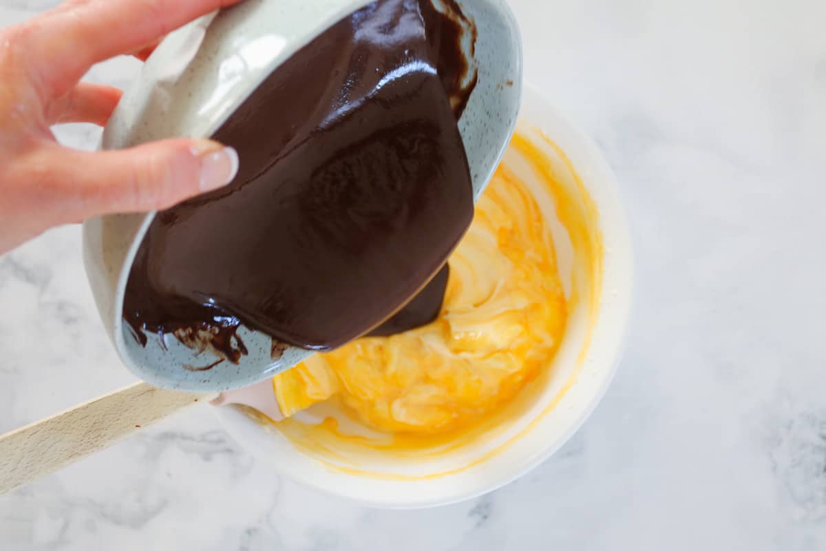 Melted chocolate being poured into a white bowl filled with cream and eggs.