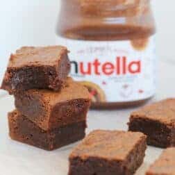 A stack of three chocolate brownies in front of a jar of chocolate hazelnut spread.