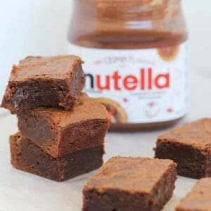 A stack of three chocolate brownies in front of a jar of chocolate hazelnut spread.