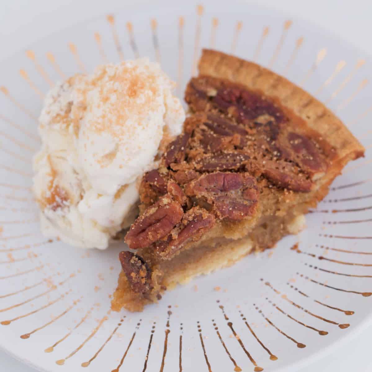 A slice of pecan tart with a scoop of vanilla ice-cream on a white and gold plate.
