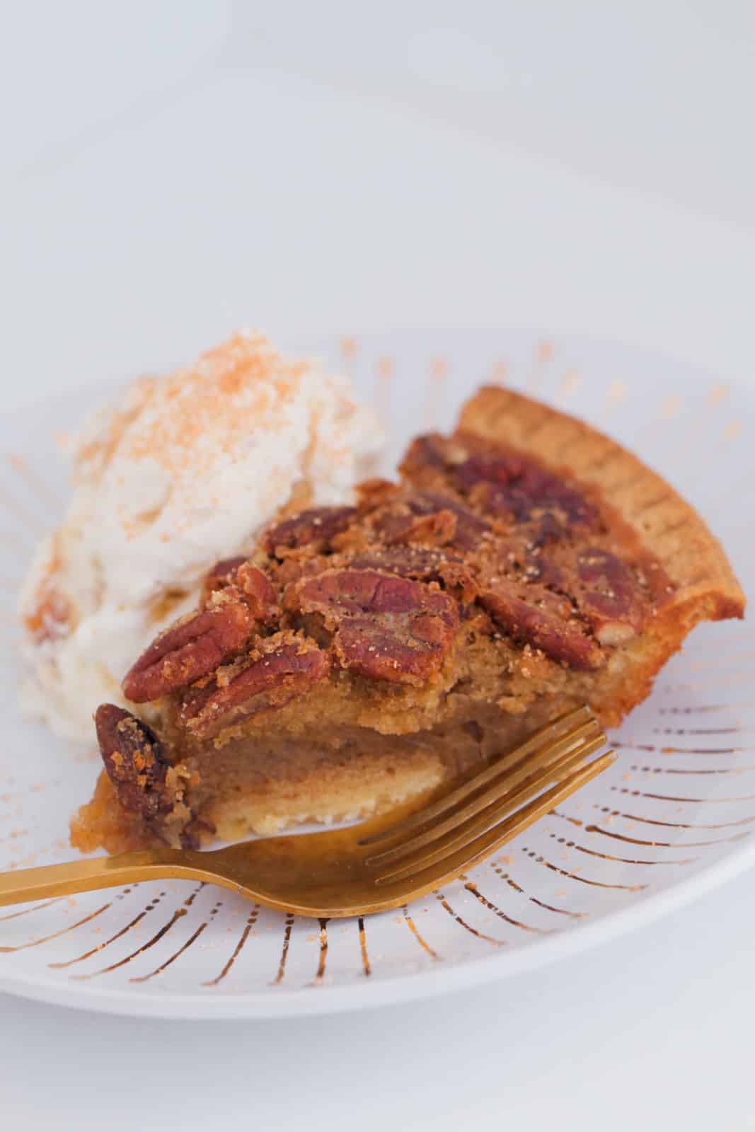 A gold fork in front of a pie of caramel pecan pie with ice-cream in the background.