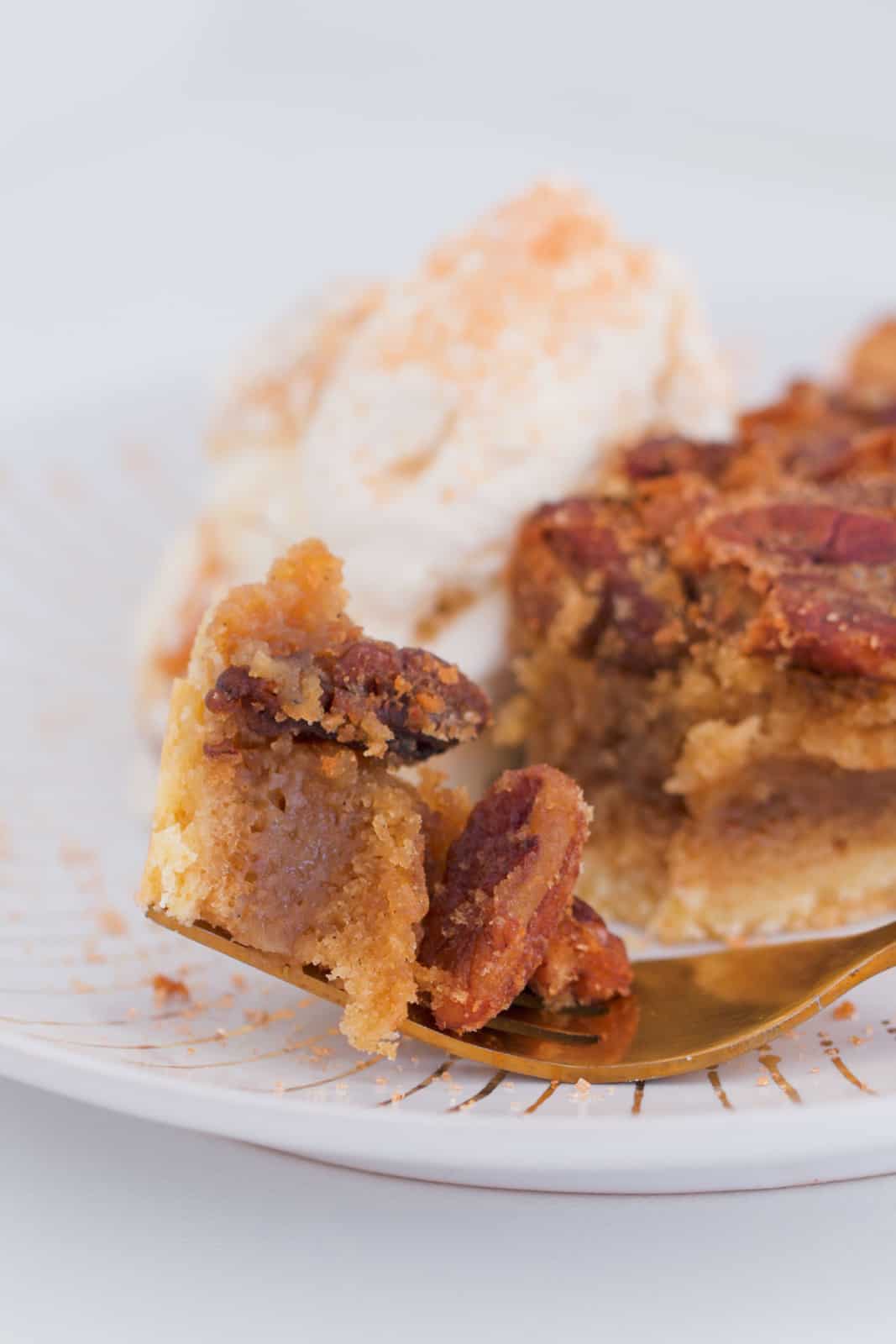 A piece of caramel pecan pie on a gold fork with ice-cream in the background.