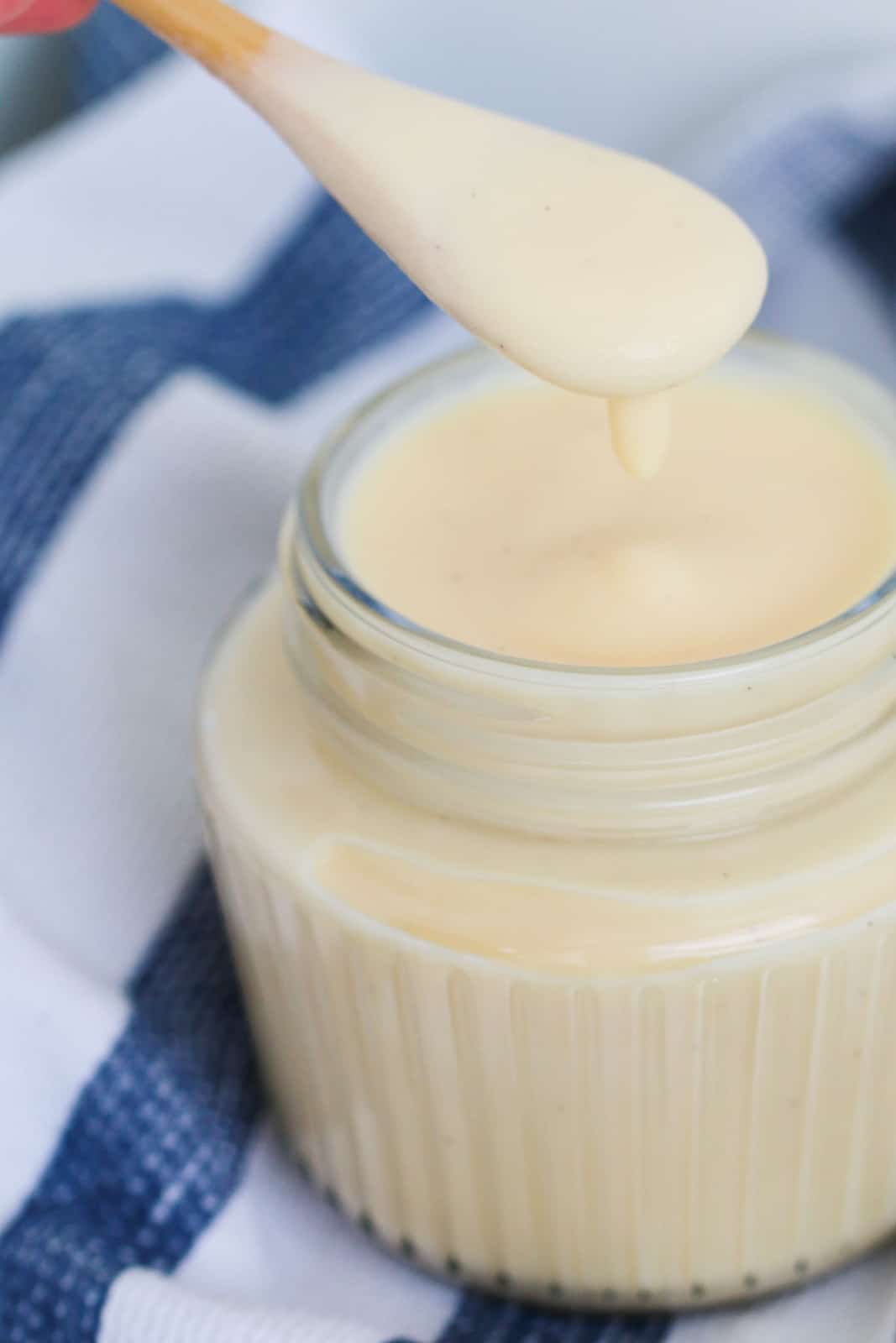 Creamy custard dripping off a wooden spoon into a glass jar of vanilla custard.
