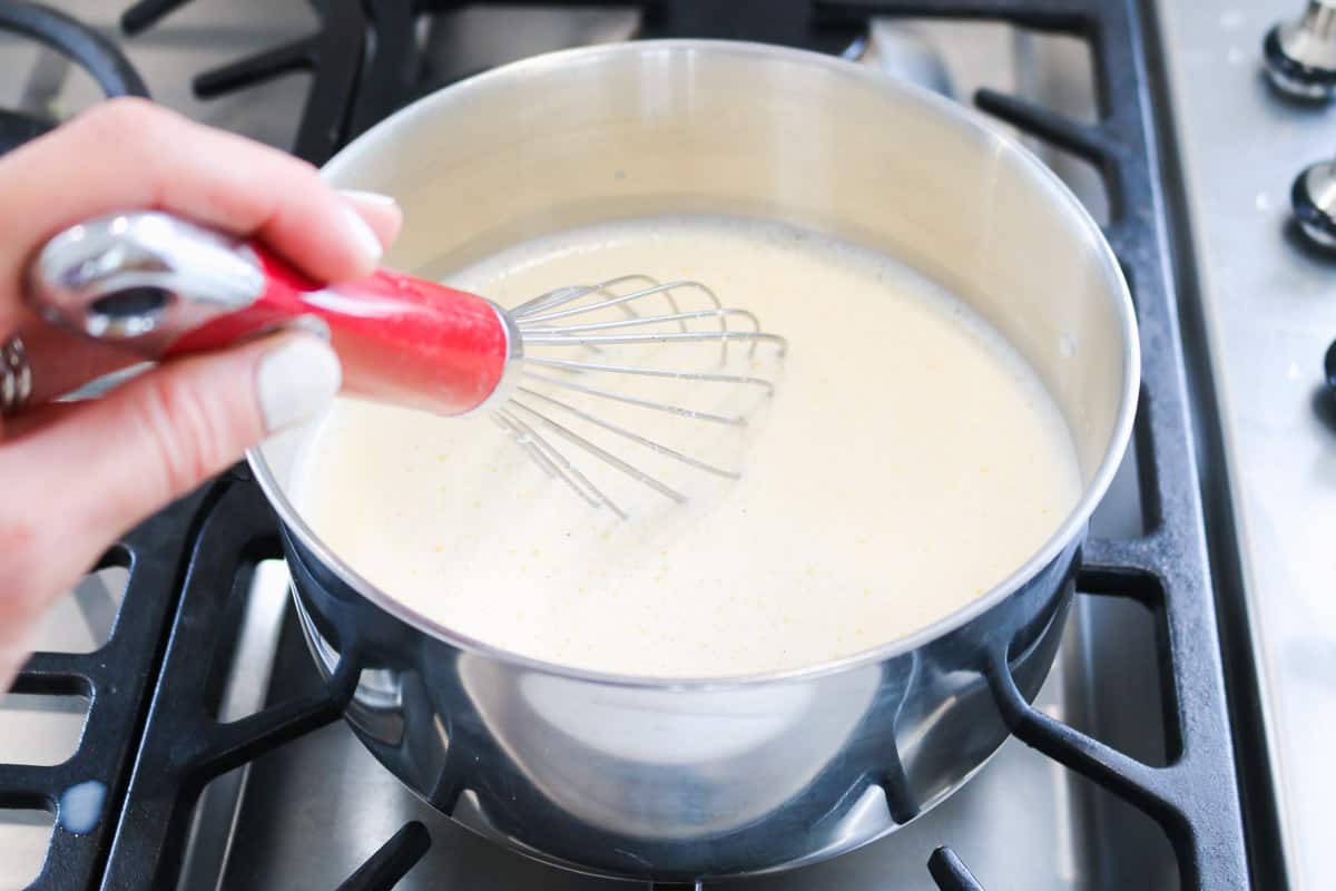 A whisk stirring a creamy liquid custard in a saucepan on a hotplate