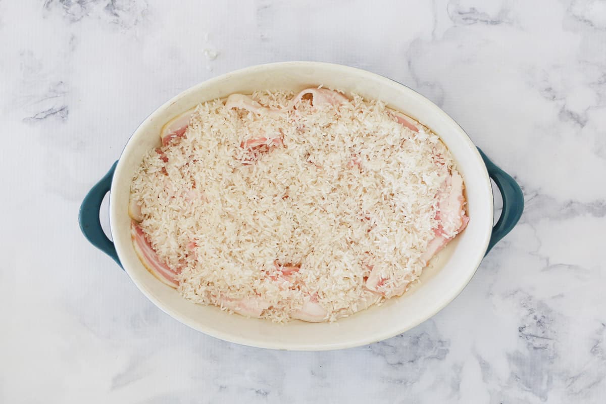 Rice sprinkled over bacon in an oval  casserole dish.