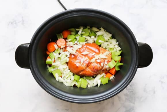 A ham hock, celery, carrot and onion on top of split peas in a slow cooker.