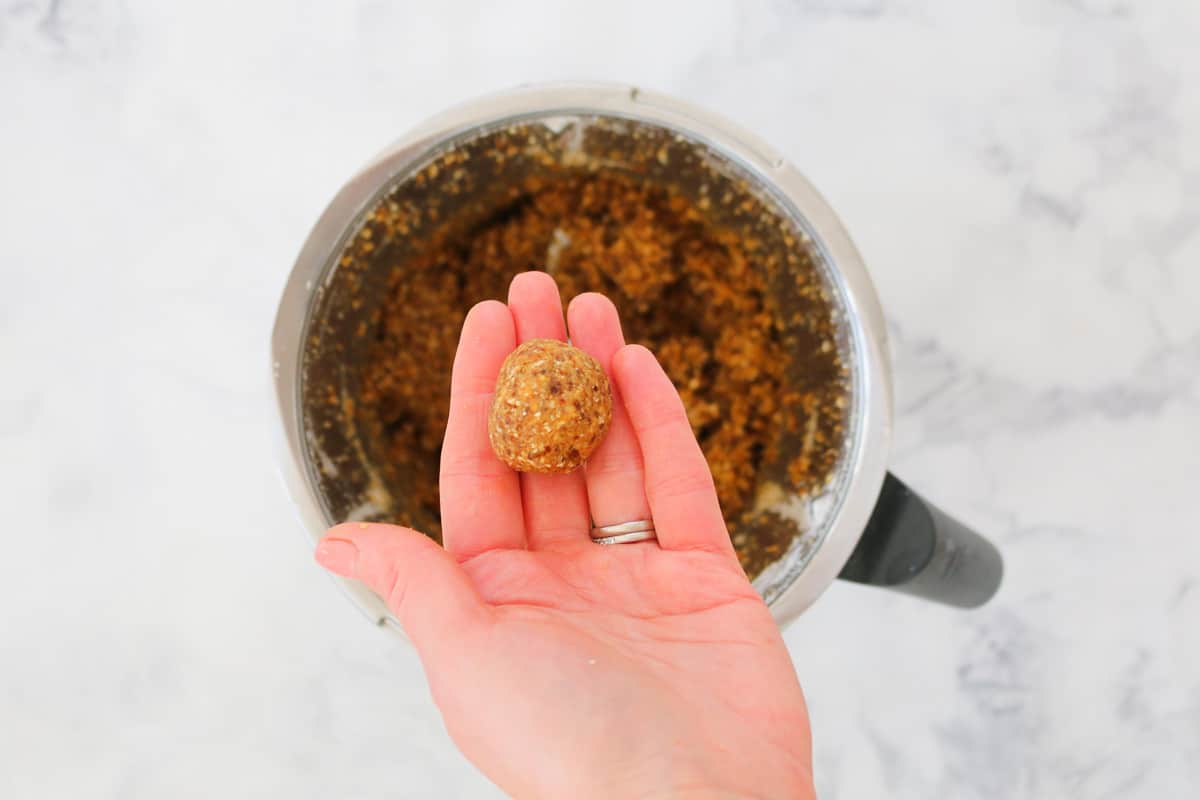 A hand with a rolled caramel protein ball over a bowl with mixed ingredients inside
