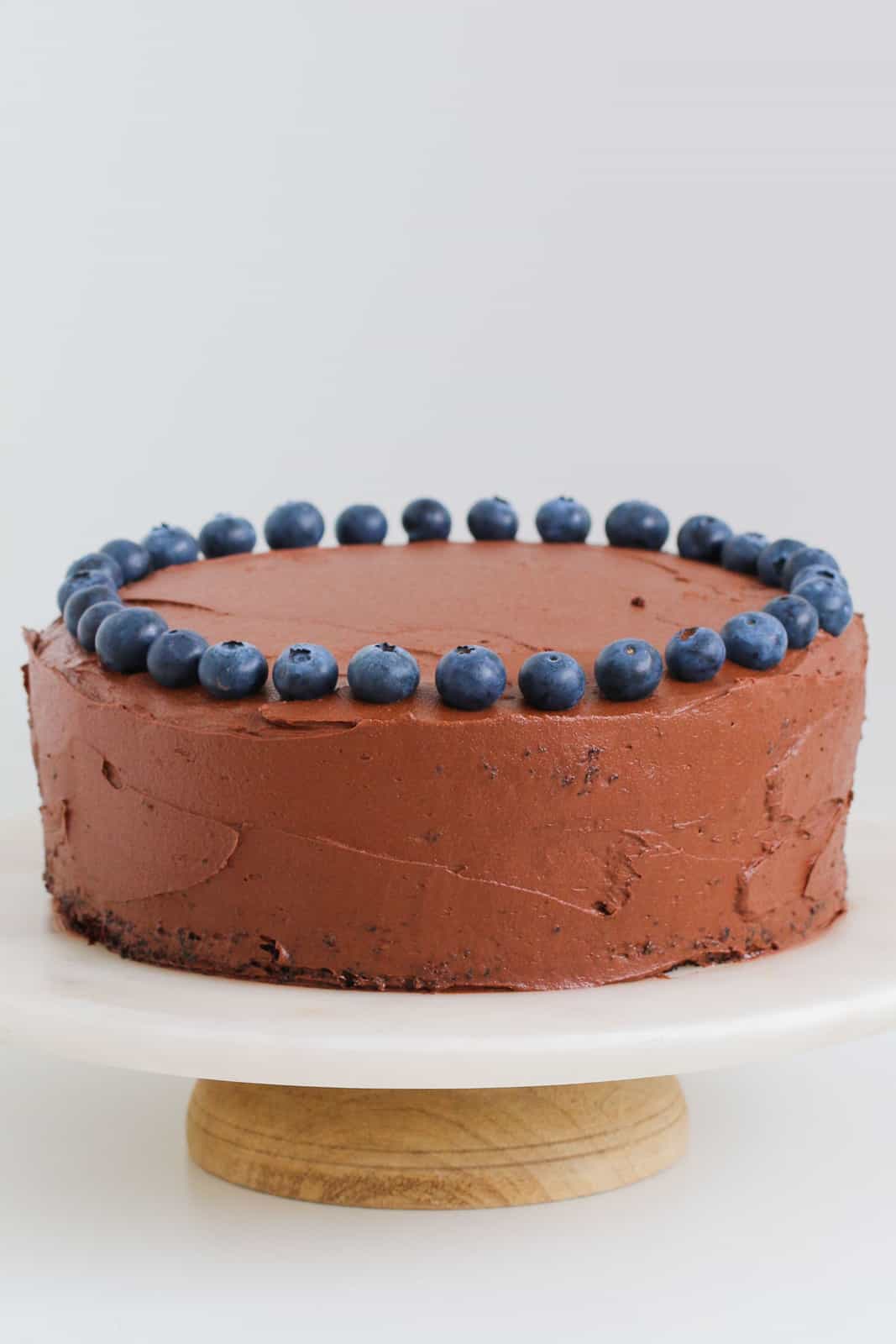A round chocolate cake on a cake stand decorated with chocolate frosting and blueberries.