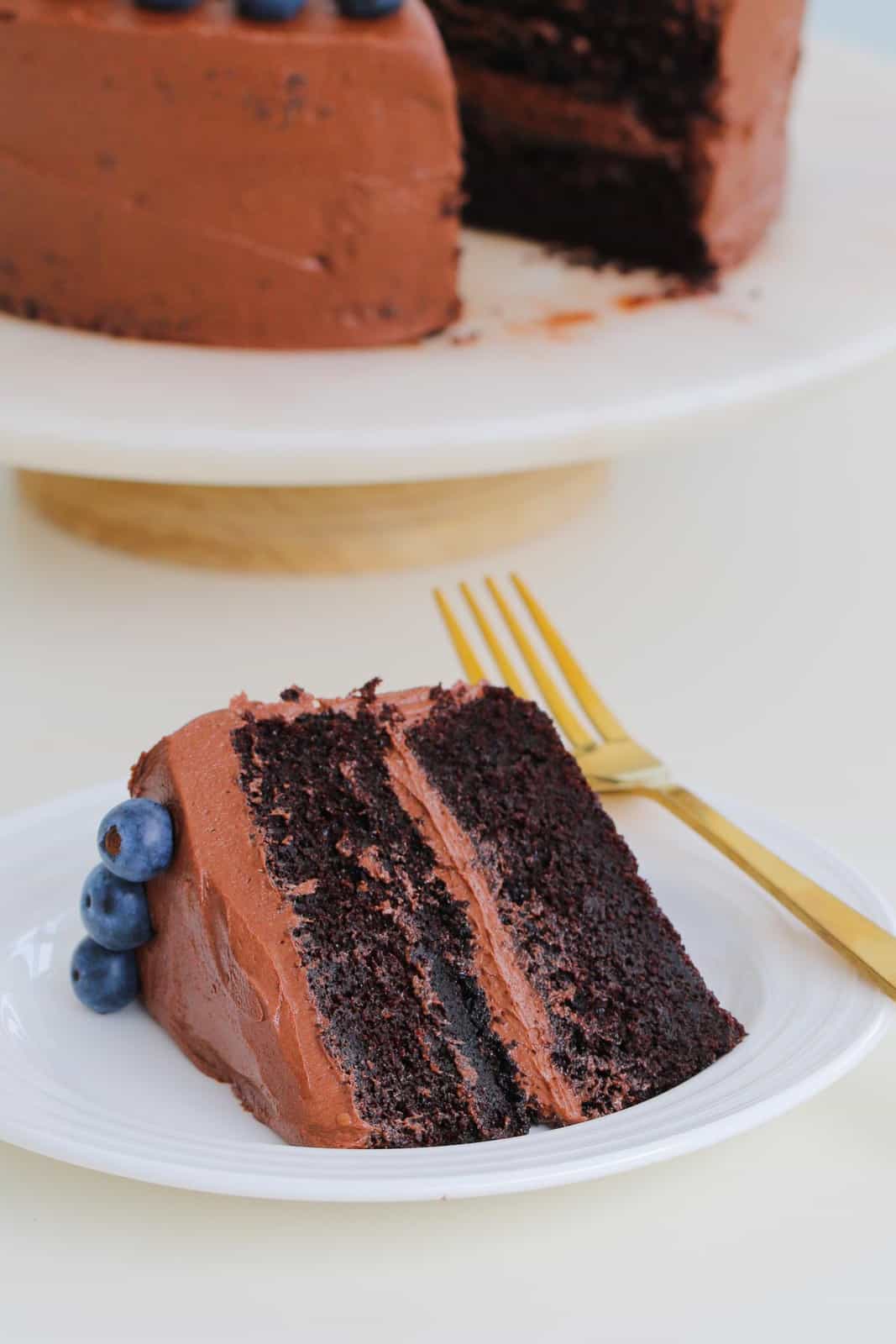 A slice of rich mud layered cake with chocolate frosting and blueberries on a white plate with a gold fork.