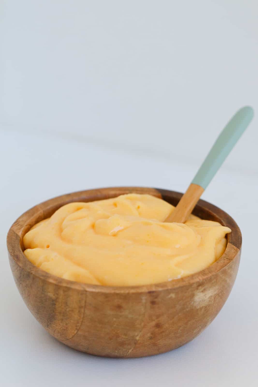 A spoon in a small wooden bowl filled with creamy yellow lemon curd.