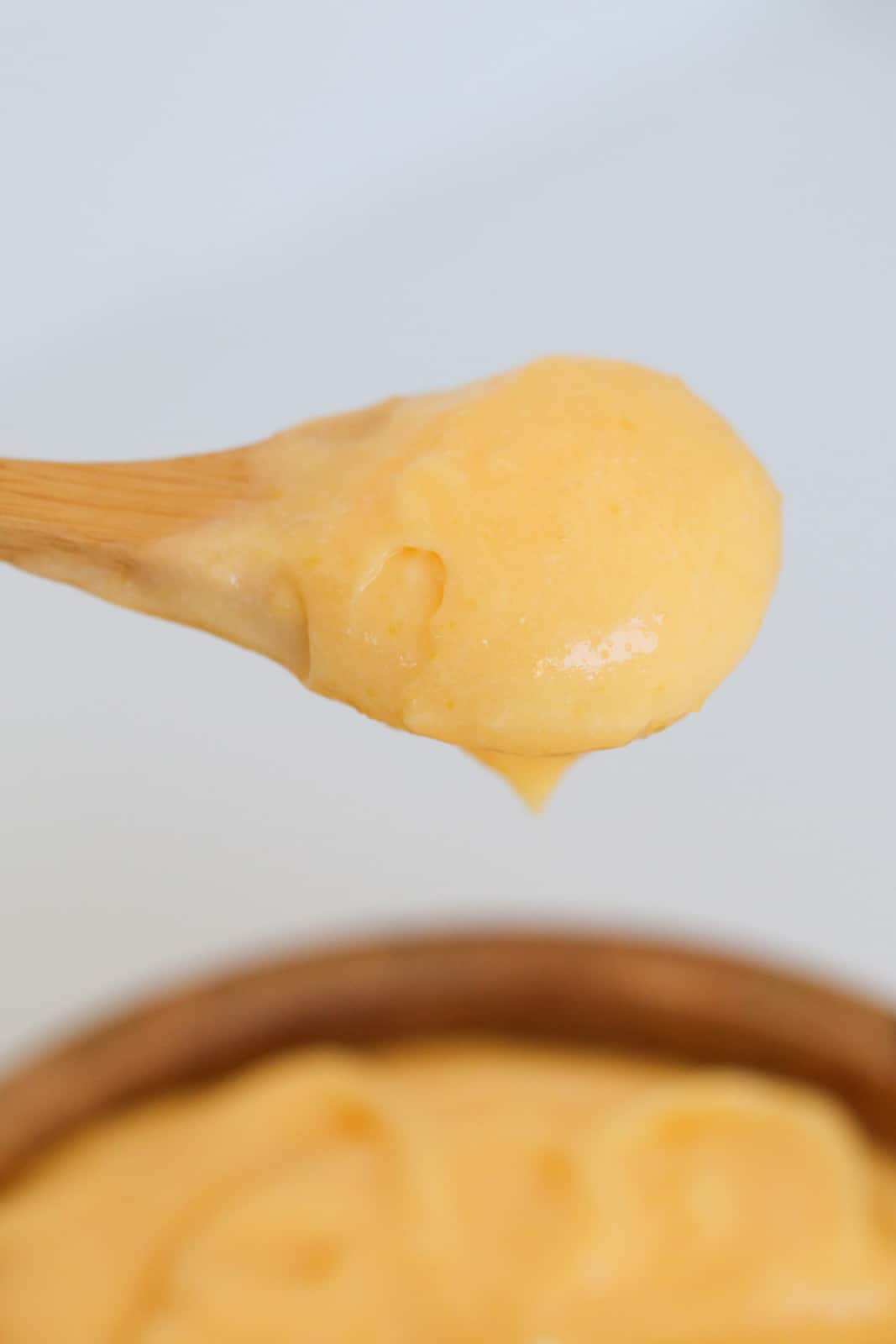 A wooden spoonful of creamy lemon butter curd held above a wooden bowl.
