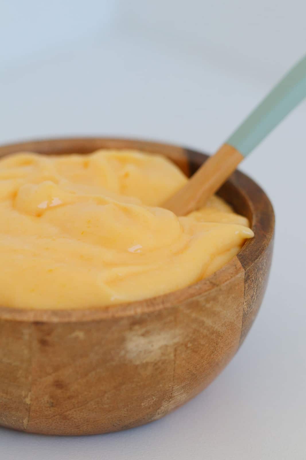 A close up of a spoon in a small wooden bowl filled with creamy yellow lemon curd.